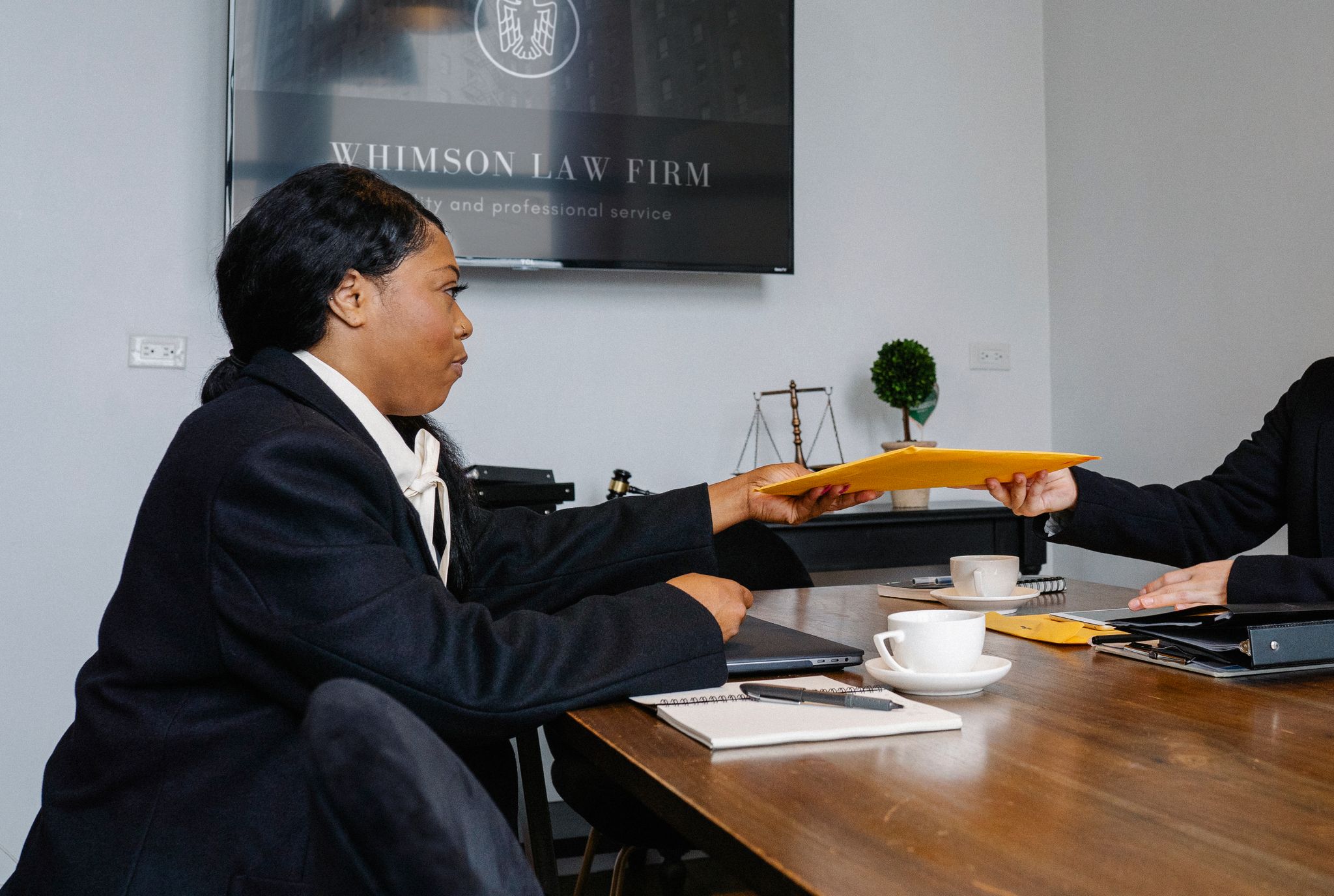 Side view of African American woman in formal wear sitting at table with documents and laptop while surrendering documents to colleague